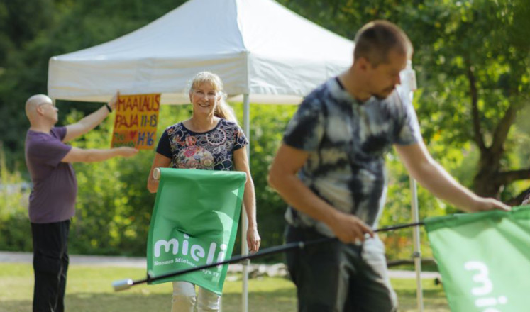 Neljän hengen hyväntuulinen ryhmä kokoaa tapahtumapistettä, viirejä ja kylttejä, telttakatoksen luona