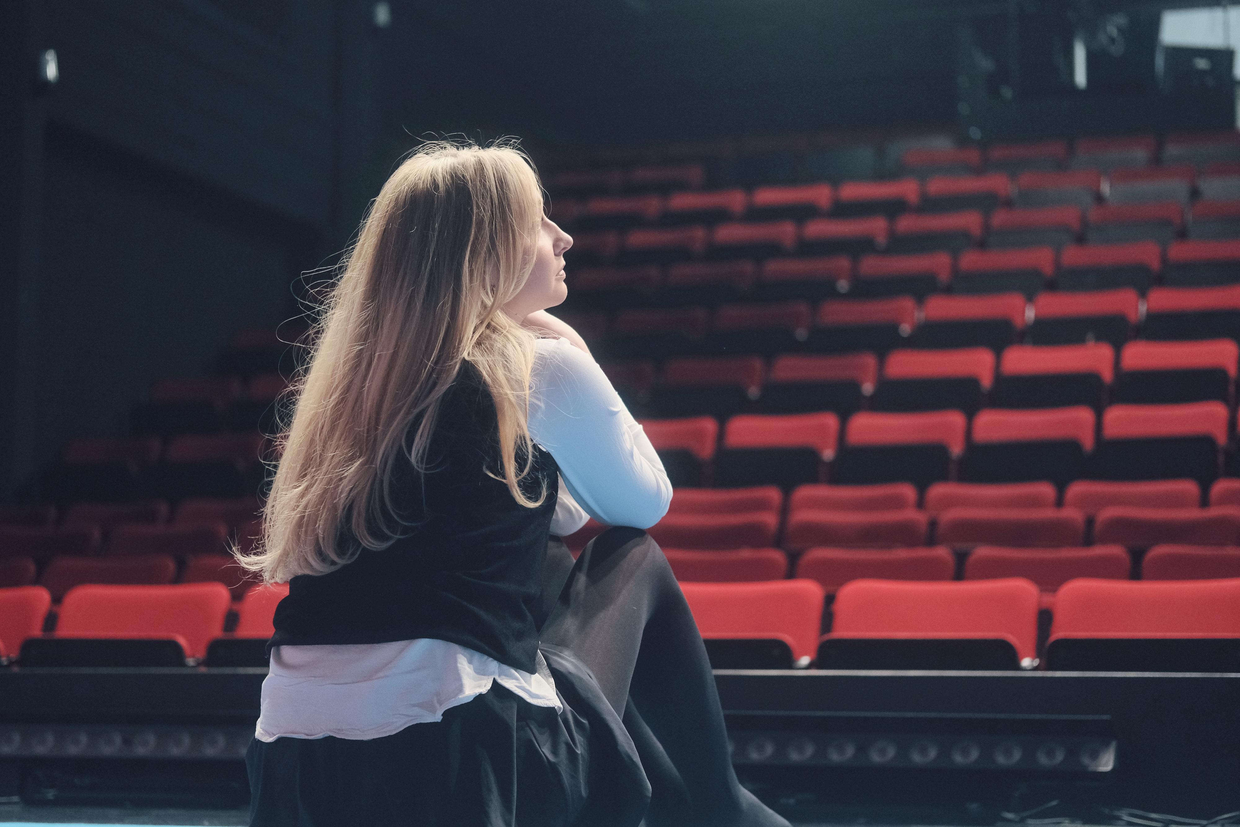 Tyttö istuu teatterisalissa, katseet kohti tyhjiä penkkiä.